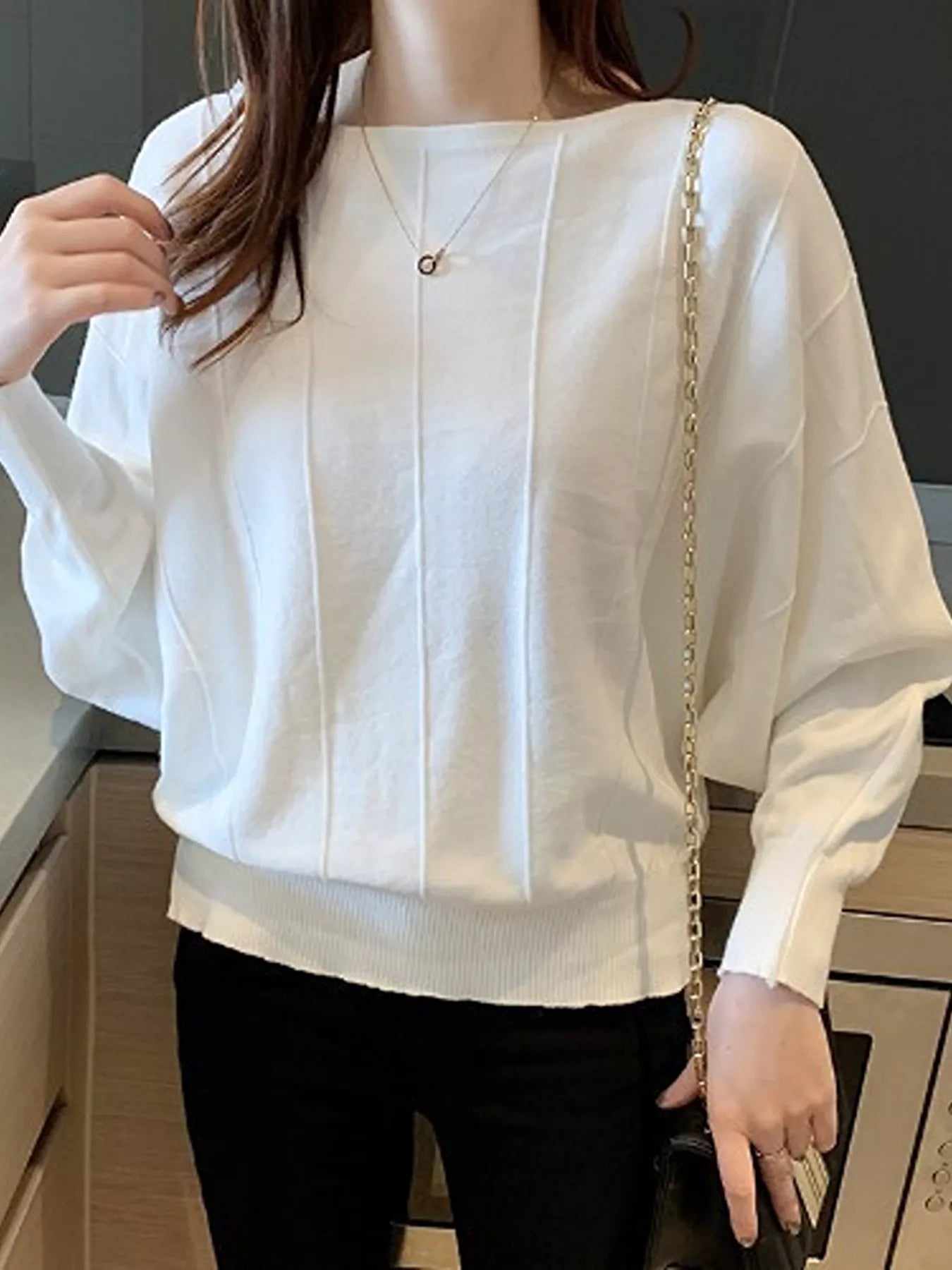 Close front view of the white batwing sleeve knit top with vertical seams, worn indoors with a chain bag.