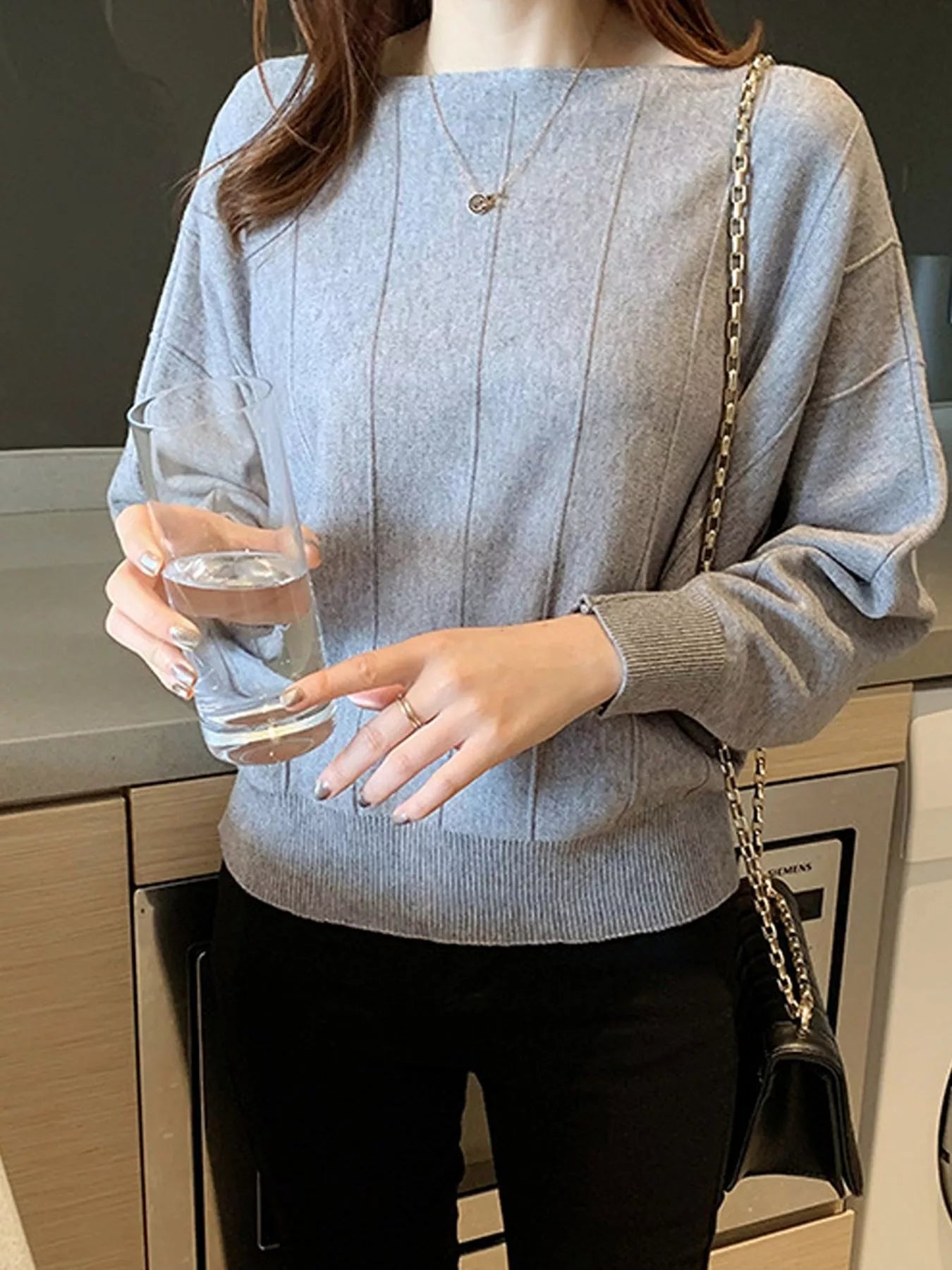 Front view of the gray batwing sleeve knit top with vertical seams, holding a glass indoors.